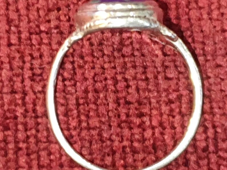 May include: A silver ring with a purple stone set in the center. The ring is on a red fabric background.