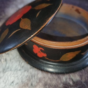 May include: A black wooden trinket box with a lid. The box is decorated with hand-painted red and gold flowers. The lid is open and the inside of the box is visible.