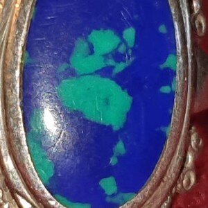 May include: A silver ring with an oval-shaped blue and green gemstone set in the center. The ring has a decorative, ornate design around the edge.