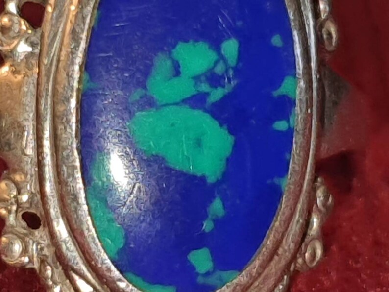 May include: A silver ring with an oval-shaped blue and green gemstone set in the center. The ring has a decorative, ornate design around the edge.