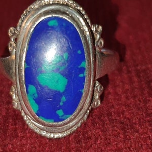May include: A silver ring with an oval-shaped blue and green gemstone set in a silver bezel. The ring has a decorative design around the bezel.
