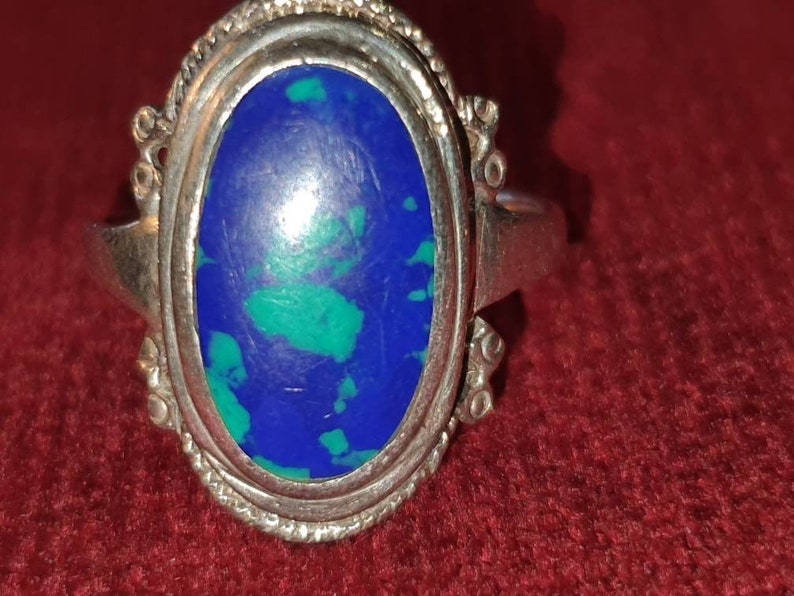 May include: A silver ring with an oval-shaped blue and green gemstone set in a silver bezel. The ring has a decorative design around the bezel.