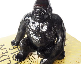 May include: A glossy black gorilla figurine in a seated position. The sculpture has a detailed muscular physique and is resting on a book with gold lettering. The figurine is a decorative item.