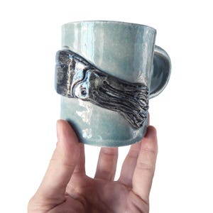 May include: A ceramic mug with a glossy light blue glaze featuring a raised, dark gray sculptural relief of a squid wrapped around the exterior. The mug has a cylindrical body and a curved handle extending from the side of the vessel.
