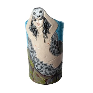 May include: A ceramic vase featuring a mermaid design. The mermaid has long black hair and is holding a seal mask. The vase is painted with blue, gray, and green colors.
