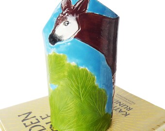 May include: A ceramic vase with a hand-painted okapi design. The vase features a blue sky background, a green leaf, and the okapi's brown and white markings. The vase is resting on a book.