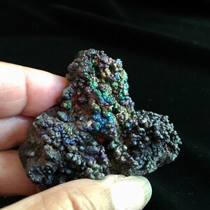 May include: A close-up of a dark gray iridescent mineral with a rough, textured surface. The mineral has a variety of colors, including blue, green, and purple.