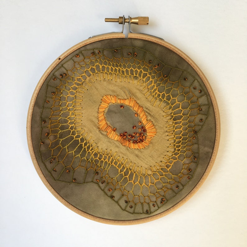 May include: A circular embroidery hoop with a green and yellow embroidered design. The design features a central circle with a brown center and orange and yellow stitching. The outer circle is a repeating pattern of hexagons.