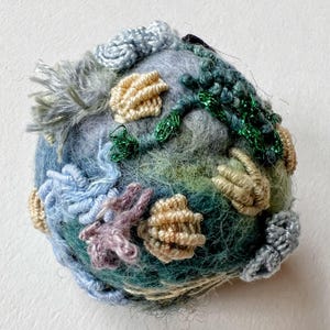 Pu&ograve; includere: Una piccola scultura rotonda in feltro con una variet&agrave; di texture e colori. La scultura &egrave; realizzata in lana e presenta una variet&agrave; di colori, tra cui blu, verde e beige. La scultura &egrave; decorata con una variet&agrave; di texture, tra cui lisce, ruvide e irregolari.