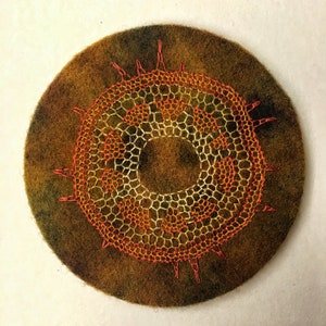May include: A circular piece of felt with a brown and green ombre effect. A large, orange and yellow embroidered design is centered on the felt. The design is a circular pattern with a ring in the center.