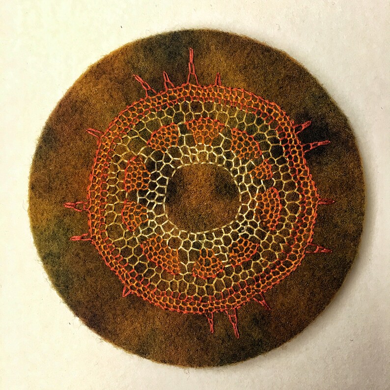 May include: A circular piece of felt with a brown and green ombre effect. A large, orange and yellow embroidered design is centered on the felt. The design is a circular pattern with a ring in the center.