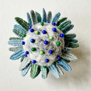 Pu&ograve; includere: Un cuscino per spilli in feltro grigio con perline blu e verdi, circondato da un anello di petali di tessuto blu e verde.