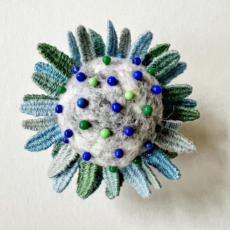 Pu&ograve; includere: Un cuscino per spilli in feltro grigio con perline blu e verdi, circondato da un anello di petali di tessuto blu e verde.