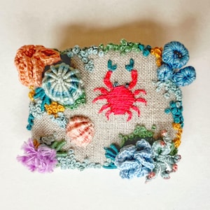 May include: A rectangular piece of light brown linen fabric featuring a central embroidered bright pink crab surrounded by textured, three-dimensional embroidery of colorful coral, sea anemones, and shells in shades of blue, orange, and purple.