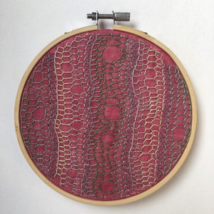 May include: A circular embroidery hoop with a pink fabric background and a detailed design of overlapping geometric shapes in shades of green, silver, and gold.