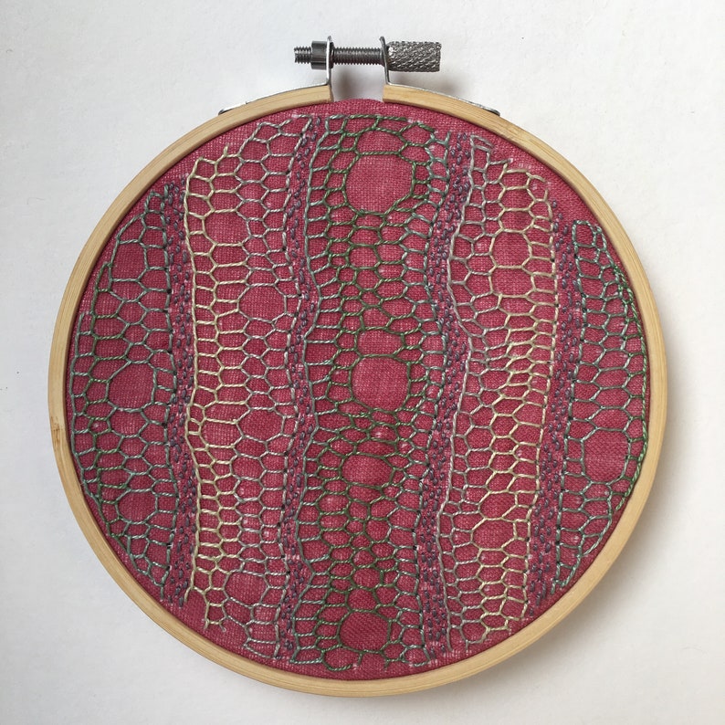 May include: A circular embroidery hoop with a pink fabric background and a detailed design of overlapping geometric shapes in shades of green, silver, and gold.