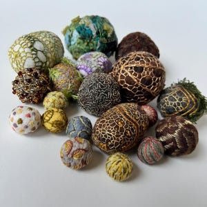 May include: A collection of small, hand-stitched spheres in various colors and textures, including brown, gold, green, white, and gray. The spheres are decorated with intricate embroidery and beading.