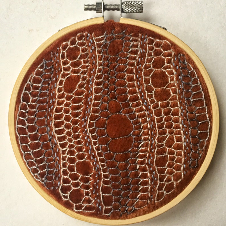 May include: A brown velvet hoop with a white and blue embroidered design. The design is a repeating pattern of circles and lines.