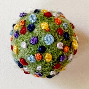 Pu&ograve; includere: Una palla di feltro verde ricoperta di fiori di feltro colorati. I fiori sono in tonalit&agrave; di rosso, arancione, giallo, blu, viola e bianco.