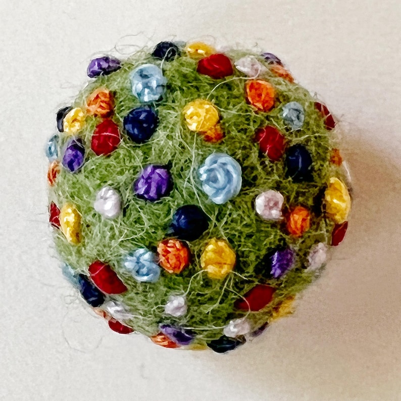 Pu&ograve; includere: Una palla di feltro verde ricoperta di fiori di feltro colorati. I fiori sono in tonalit&agrave; di rosso, arancione, giallo, blu, viola e bianco.