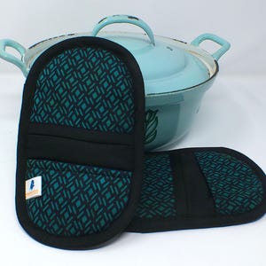 Könnte beinhalten: Zwei schwarze und tealfarbene Ofenhandschuhe mit geometrischem Muster, neben einem hellblauen Topf mit Griffen. Die Handschuhe haben eine schwarze Umrandung und ein schwarzes Band in der Mitte. Ein kleines Etikett ist auf einem Handschuh sichtbar.