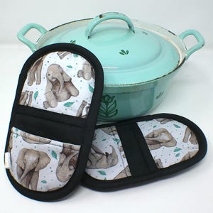 Może przedstawiać: Dwa czarne rękawice kuchenne z nadrukiem słonia i jasnoniebieski garnek z pokrywką. Rękawice mają białe tło z szarymi słoniami i zielonymi liśćmi. Garnek jest jasnoniebieski z uchwytem po każdej stronie.