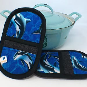 Könnte beinhalten: Zwei schwarze und blaue Ofenhandschuhe und ein passender Topflappen mit Waldesign. Die Handschuhe und der Topflappen haben einen schwarzen Rand und einen blauen Hintergrund mit weißen und schwarzen Orcas. Im Hintergrund ist ein hellblauer Topf.