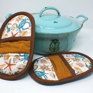 Könnte beinhalten: Zwei braune und weiße Topflappen mit einem Muschel- und Krabbenmuster. Die Topflappen haben einen braunen Stoffeinsatz. Ein hellblauer emaillierter Schmortopf mit grünem Blumendesign ist im Hintergrund.