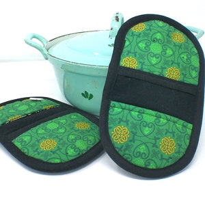 Könnte beinhalten: Zwei Topflappen in ovaler Form mit grünem und goldenem Muster und schwarzem Besatz. Die Topflappen haben auf einer Seite eine Tasche.