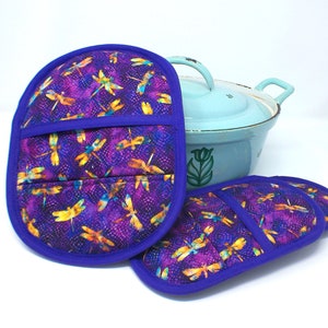 Könnte beinhalten: Zwei lila Topflappen mit einem Libellenmuster und blauem Besatz. Die Topflappen sind aus Stoff und haben eine Tasche auf der Vorderseite.