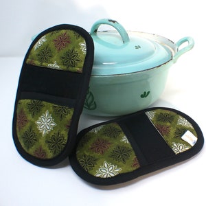Könnte beinhalten: Zwei grüne und schwarze Topflappen mit Blumenmuster. Die Topflappen sind oval geformt und haben eine schwarze Stoffmitte.