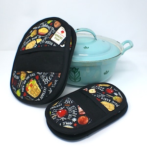 May include: Two black oven mitts with a colorful apple and fall-themed design. The mitts have a pocket on the back and feature text like "The Great Apple Pie", "Home Baked", and "Crazy for Candy Apples".