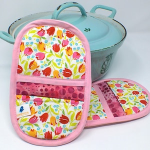 Könnte beinhalten: Zwei rosa Ofenhandschuhe mit einem floralen Tulpenmuster und einem hellblauen Topf im Hintergrund. Die Handschuhe haben eine rosa Bordüre und eine Tasche mit einem Tupfenmuster. Das Tulpenmuster umfasst rote, gelbe und rosa Blumen.