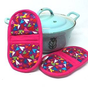 May include: Two pink oven mitts with a colorful heart pattern. The mitts are made of fabric and have a quilted lining.