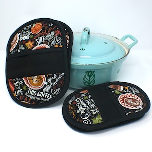 May include: Three black oven mitts with a fall-themed coffee and pumpkin design. The mitts have a black pocket in the middle and are printed with text such as "This coffee is making me awesome" and "Good morning coffee".