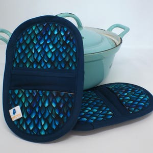 Könnte beinhalten: Zwei marineblaue Topflappen mit einem blau-türkisfarbenen Schuppenmuster, neben einem hellblauen Topf. Die Topflappen haben ein Taschen-Design und ein Etikett auf einem. Der Topf hat zwei Griffe und einen Deckel.