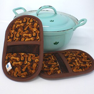 Könnte beinhalten: Zwei braune Topflappen mit einem Knochenmuster. Die Topflappen sind aus Stoff und haben einen braunen Rand. Ein hellblauer Emailletopf ist im Hintergrund.