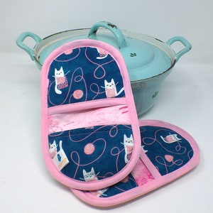 Könnte beinhalten: Ein Paar rosa und blaue Topflappen mit Katzenmuster. Die Topflappen haben eine Tasche auf der Rückseite und sind mit weißen Katzen verziert, die Wollknäuel halten.