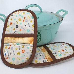Könnte beinhalten: Zwei braune und weiße Topflappen mit einem Pfoten- und Knochenmuster. Die Topflappen haben einen braunen Rand und einen beigefarbenen Stoffeinsatz. Die Topflappen liegen neben einem hellblauen Emailletopf mit Deckel.