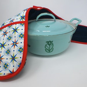 May include: A light blue enamel Dutch oven with a matching lid and handles, paired with a patterned oven mitt and pot holder. The mitt features a white background with red, blue, and yellow starburst designs. The pot holder is navy blue.