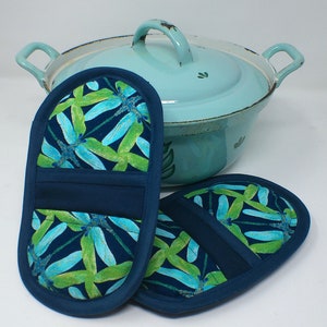 Puede incluir: Un par de agarraderas de olla azules con un patrón de libélula. Las agarraderas de olla están hechas de tela y tienen un borde azul oscuro. Las agarraderas de olla están sobre una superficie blanca junto a una olla azul claro con tapa.
