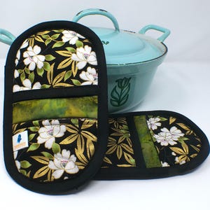 Könnte beinhalten: Schwarze und goldfarbene Ofenhandschuhe mit weißen Blumen und grünen Akzenten. Im Hintergrund ein hellblauer Kochtopf. Die Handschuhe haben einen schwarzen Rand und einen grünen Stoffakzent.