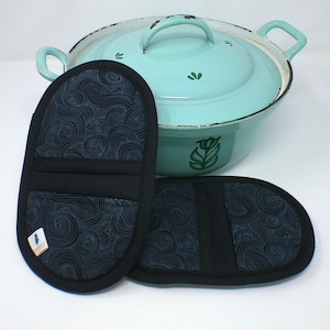 Könnte beinhalten: Ein türkisfarbener Emaille-Topf mit Deckel und zwei schwarzen Topflappen. Der Topf hat einen weißen Rand und grüne Blumendesigns. Die Topflappen haben ein schwarzes Wellenmuster und einen schwarzen Rand.