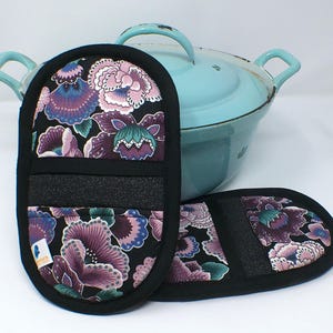 Könnte beinhalten: Zwei geblümte Ofenhandschuhe mit schwarzem Rand und schwarzem Handband. Die Handschuhe haben einen schwarzen Hintergrund mit lila, rosa und türkisfarbenen Blumen. Im Hintergrund befindet sich ein hellblauer Topf mit Deckel und Griffen.