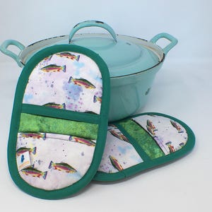 Könnte beinhalten: Zwei grün umrandete Topflappen mit Fischmuster neben einem hellblauen Kochtopf. Die Topflappen haben einen weißen Hintergrund mit Aquarell-Fischillustrationen und einem grünen Streifen. Der Topf hat einen passenden hellblauen Deckel und Griffe.