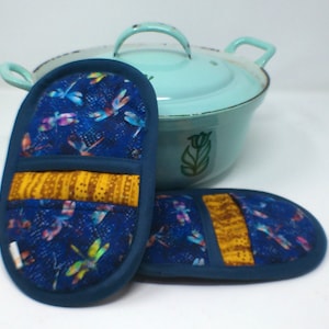 Könnte beinhalten: Zwei blau-gelbe, gesteppte Topflappen mit Libellenmuster. Die Topflappen sind oval geformt mit dunkelblauem Rand und gelbem Band. Im Hintergrund ein hellblauer Emaille-Topf mit Deckel.