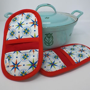 May include: Two red and white oven mitts with a compass rose pattern, next to a light blue Dutch oven. The mitts have a red trim and a red band across the middle. The Dutch oven has a matching light blue lid.