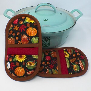 May include: Two oven mitts and a teal pot. The mitts feature a black background with a fall-themed pattern of pumpkins, apples, and leaves. The mitts have a brown border and a red center. The pot is teal with white accents.