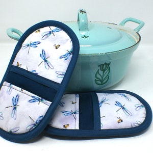 Op de afbeelding: Twee blauwe en witte ovenwanten met een libellen- en bijenpatroon. De ovenwanten liggen op een wit oppervlak naast een lichtblauwe emaillepan met een groen tulpenmotief.
