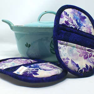 Puede incluir: Dos agarraderas para ollas de color blanco y morado con ribete azul. Las agarraderas están hechas de tela y tienen un diseño acolchado. Las agarraderas están sobre una superficie blanca junto a una olla azul claro con tapa.